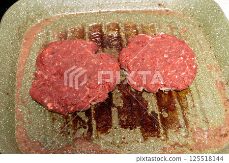Two raw burger patties on a speckled green grill pan, with grease marks emphasizing the homemade preparation process before grilling. 125518144