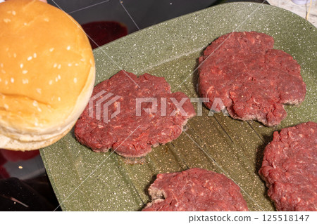 Raw hamburger patties on a green grill pan with a sesame seed bun, capturing the simplicity of preparing fresh, homemade burgers. 125518147