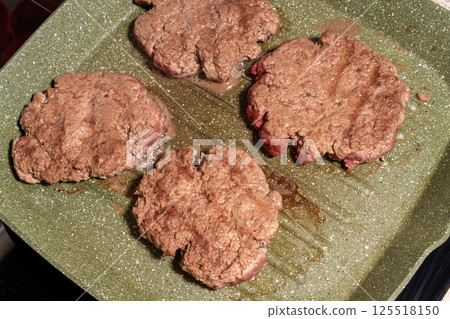 Four sizzling hamburger patties cook on a green griddle, showing texture, grease, and browning. A snapshot of burger preparation at its finest. 125518150