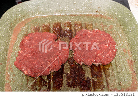Two raw burger patties on a speckled green grill pan, with grease marks emphasizing the homemade preparation process before grilling. 125518151