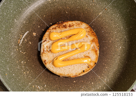 A sesame seed bun with zigzag mustard lines rests on a skillet, surrounded by crumbs and sesame seeds, ready for burger assembly. 125518153