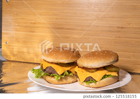 Two cheeseburgers with beef patties, cheddar cheese, pickles, lettuce, and tomatoes on sesame buns, served on a white plate with a rustic background. Two cheeseburgers with beef patties, cheddar cheese, pickles, lettuce, and tomatoes on sesame buns, served on a white plate with a rustic background. 125518155