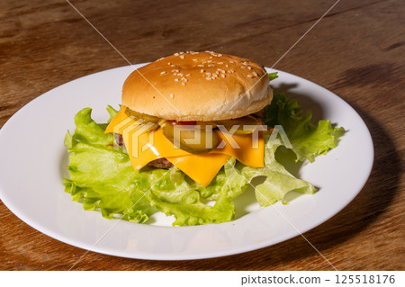 A juicy cheeseburger with a sesame seed bun, melted cheese, beef patty, and lettuce on a white plate, styled atop a rustic wooden background. A juicy cheeseburger with a sesame seed bun, melted cheese, beef patty, and lettuce on a white plate, styled atop a rustic wooden background. 125518176