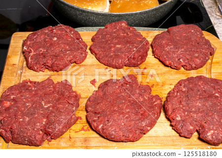 Uncooked burger patties on a wooden cutting board with hamburger buns being toasted in the background. Perfect for BBQ or homemade cooking scenes. Uncooked burger patties on a wooden cutting board with hamburger buns being toasted in the background. Perfect for BBQ or homemade cooking scenes. 125518180