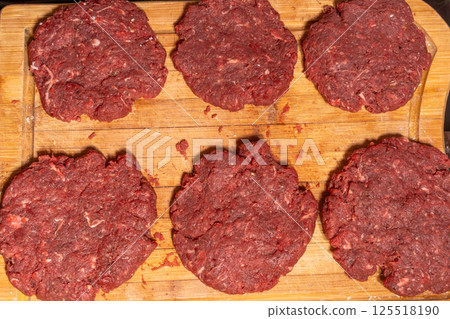Uncooked burger patties on a wooden cutting board with hamburger buns being toasted in the background. Perfect for BBQ or homemade cooking scenes. 125518190
