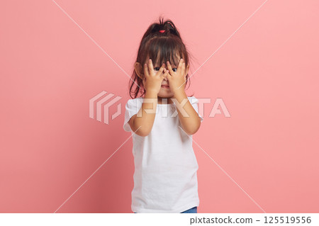 Asian little girl looks through her fingers, covers her face with both hands, scared expression, as she notices something scary or frightening, isolated on a pink background. 125519556