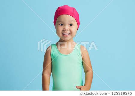 Little cheerful girl swimmer wears pink cap and swimming costume, prepares for workout in pool, smiles broadly, stands over blue background with blank space Little cheerful girl swimmer wears pink cap and swimming costume, prepares for workout in pool, smiles broadly, stands over blue background with blank space 125519570