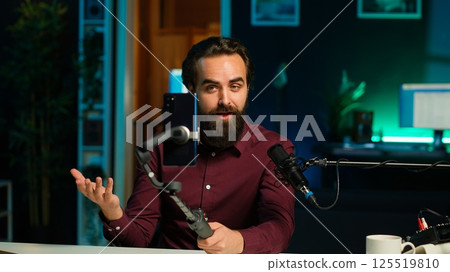 Expressive man using smartphone on selfie stick to shoot video, doing frantic gesturing. Animated influencer making hectic hand movements while filming vlog with phone in studio, camera A 125519810