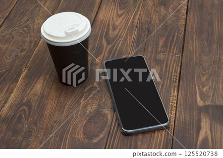 A smartphone with an unused screen and a paper coffee cup on a wooden table, ideal for a quick break during work. A smartphone with an unused screen and a paper coffee cup on a wooden table, ideal for a quick break during work. 125520738
