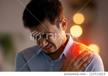A man is clutching his shoulder in pain with a grimace, highlighted by red light to indicate the affected area of discomfort. 125521623