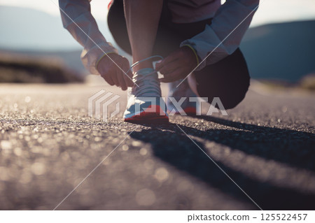 Woman runner tying shoelace on sunset mountain road Woman runner tying shoelace on sunset mountain road 125522457