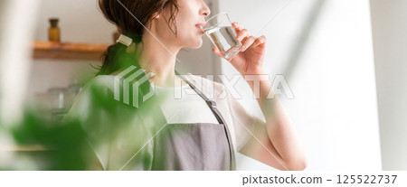 A woman in an apron drinking water from a cup in the kitchen (hydration, mineral water, water server) 125522737