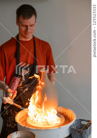 A potter burns a jug with a gas burner on a potter's wheel. Vertical photo. A potter burns a jug with a gas burner on a potter's wheel. Vertical photo. 125523333