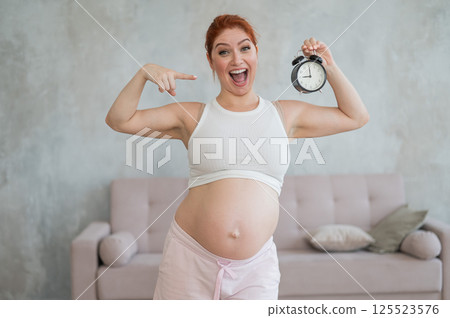 Caucasian pregnant woman holding alarm clock waiting for delivery.  125523576
