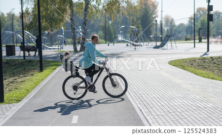 Caucasian woman riding bicycle with toddler son sitting behind her in child seat in park. Caucasian woman riding bicycle with toddler son sitting behind her in child seat in park. 125524383