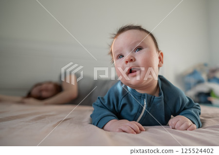 A three-month-old boy lies on his stomach on the bed and his mother sleeps behind him. Postpartum depression. 125524402