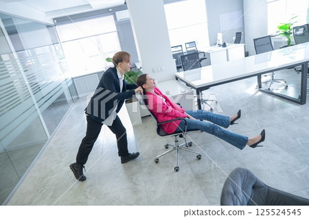 Coworkers fooling around in the office. A guy pushes a young woman around on a chair.  125524545