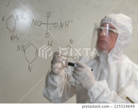 An elderly Caucasian male scientist in a protective suit holds a test tube and writes a formula on the glass.  125524649