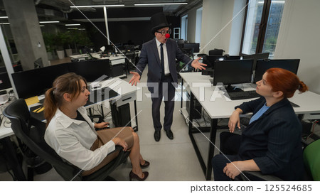 An elderly man in a clown costume fools around at work next to two Caucasian women. An elderly man in a clown costume fools around at work next to two Caucasian women. 125524685