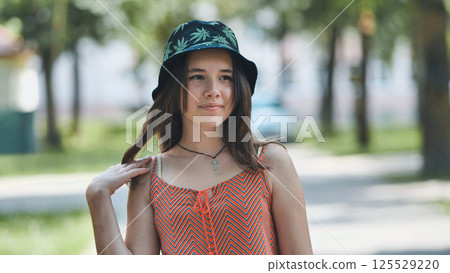 Teenage girl smiling, sporting long brown hair, bucket hat, cross necklace, relaxing during bright summer park afternoon Teenage girl smiling, sporting long brown hair, bucket hat, cross necklace, relaxing during bright summer park afternoon 125529220