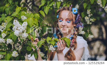 Woman wearing vibrant makeup, butterfly hair clips, inhaling fragrant white lilacs amid blossoming garden spring scenery Woman wearing vibrant makeup, butterfly hair clips, inhaling fragrant white lilacs amid blossoming garden spring scenery 125529264