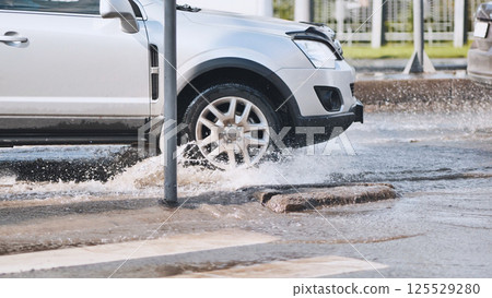 Silver sedan splashing water while driving through urban street puddle, wet asphalt reflecting vehicle's dynamic motion 125529280
