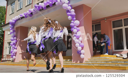 Happy graduates celebrating their last day of school, running out of the school building with purple and white balloons 125529304