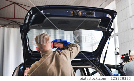 Car wash employee cleaning rear window of vehicle with open trunk 125529353