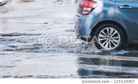 Gray car splashing through flooded city street after heavy rain, creating waves and spray 125529367