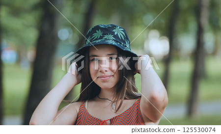 Young female wearing marijuana patterned bucket hat, relaxing during sunny park afternoon 125530074