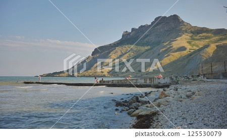 Tourists enjoying the summer sun and walking on a pier at koktebel beach in crimea, with a mountain in the background. 125530079