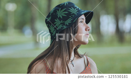 Teenage girl sporting cannabis patterned bucket hat, relaxing outdoors during sunny summer afternoon Teenage girl sporting cannabis patterned bucket hat, relaxing outdoors during sunny summer afternoon 125530181