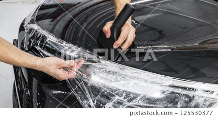 Car wrapping specialist putting vinyl foil or film on car. Protective film on the car. Applying a protective film to the car with tools. Car detailing. Transparent film. Car paint protection. Trimming Car wrapping specialist putting vinyl foil or film on car. Protective film on the car. Applying a protective film to the car with tools. Car detailing. Transparent film. Car paint protection. Trimming 125530377