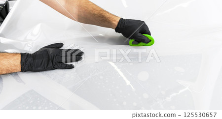 Car wrapping specialist putting vinyl foil or film on car. Protective film on the car. Applying a protective film to the car with tools. Car detailing. Transparent film. Car paint protection. Trimming 125530657