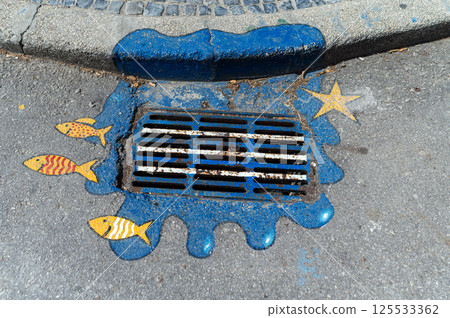 Creative artwork of a colorful fish-themed drainage grate in a city street 125533362