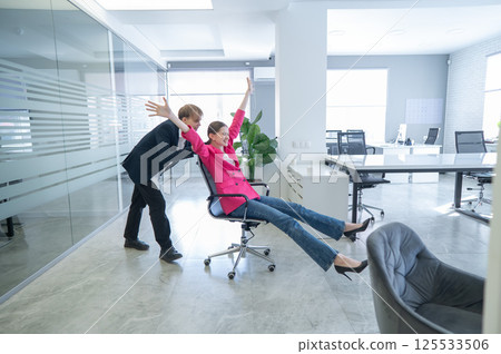 Coworkers fooling around in the office. A guy pushes a young woman around on a chair.  125533506