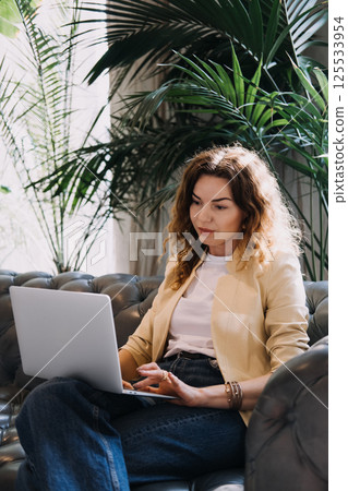 Young woman working on a laptop in a lush indoor space filled with natural light and plants. Biophilic design, wellness at work, productive environment, stress reduction... 125533954