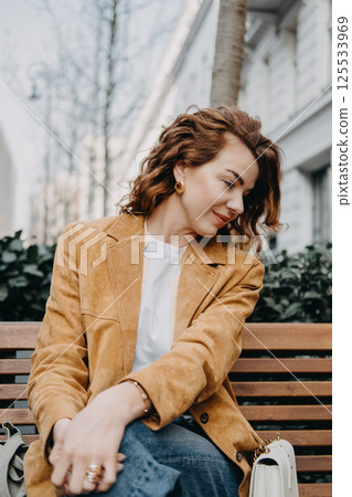 A young woman with curly red hair sits quietly on a bench looking down with a calm expression. Quiet contemplation, thoughtful mood, inner calm, soft elegance... A young woman with curly red hair sits quietly on a bench looking down with a calm expression. Quiet contemplation, thoughtful mood, inner calm, soft elegance... 125533969