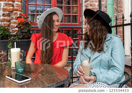two cheerful charming young women talking and drinking coffee in outdoor cafe two cheerful charming young women talking and drinking coffee in outdoor cafe 125534433
