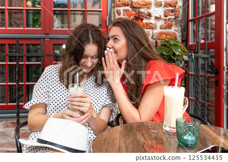 two young girls share a secret in the ear sitting in a cafe, girls party concept two young girls share a secret in the ear sitting in a cafe, girls party concept 125534435