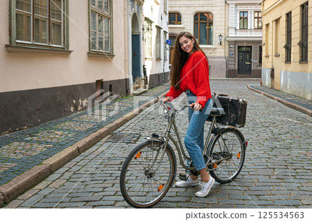 beautiful brunette with long hair in a red sweater and jeans on a vintage bike in the old town 125534563