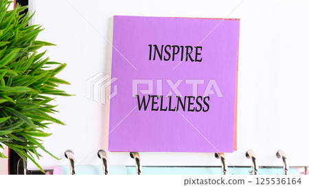 A vibrant piece of stationery features the words inspire wellness prominently. This motivational note is displayed alongside a small green plant, adding a refreshing touch A vibrant piece of stationery features the words inspire wellness prominently. This motivational note is displayed alongside a small green plant, adding a refreshing touch 125536164