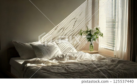 cozy bedroom scene with natural light streaming through blinds, casting shadows on messy bed with white sheets and vase of flowers on bedside table 125537440