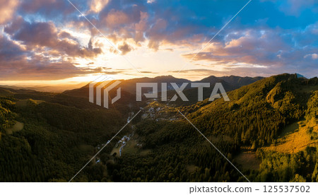 breathtaking panoramic drone shot captures mountain range during sunset, with vibrant sunbeams breaking through clouds, illuminating lush green landscape below 125537502