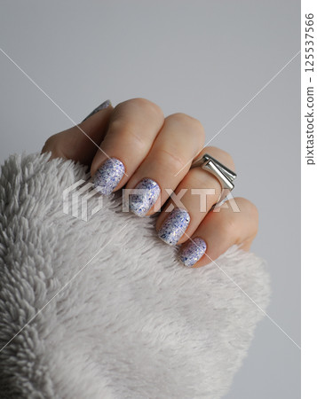 Stylish close-up of white crelly nail polish with blue shimmer, aurora flakes adorned with black foil pieces featuring silver ring and sleeve of light gray fleece sweater in neutral palette 125537566