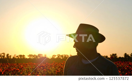 silhouette of man in hat standing in blooming poppy field, gazing downward as sun sets, creating serene and reflective atmosphere silhouette of man in hat standing in blooming poppy field, gazing downward as sun sets, creating serene and reflective atmosphere 125537872
