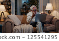 Senior man working laptop home office cozy living room remote work retirement lifestyle digital technology elderly learning comfortable environment evening warm light relaxed 125541295