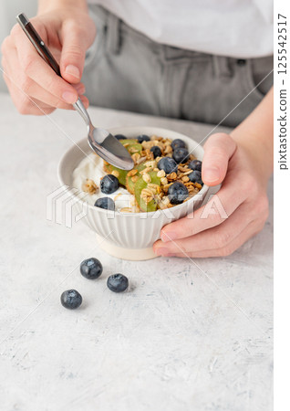 Bowl of granola with greek yogurt, kiwi and blueberries and spoon in hands of woman on white table. Bowl of granola with greek yogurt, kiwi and blueberries and spoon in hands of woman on white table. 125542517