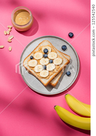 Flat lay of peanut butter toast with banana, blueberry and nuts on a plate on a pink background 125542540