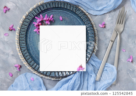 Wedding table place with a card near fabric and wild purple flowers top view on gray, spring mockup 125542845
